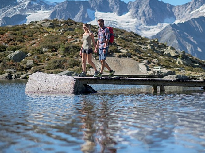 Escursioni in Alto Adige: esperienze indimenticabili! Coppia cammina su ponte sopra lago di montagna con montagne sullo sfondo