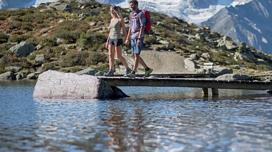Escursioni in Alto Adige: esperienze indimenticabili! Coppia cammina su ponte sopra lago di montagna con montagne sullo sfondo