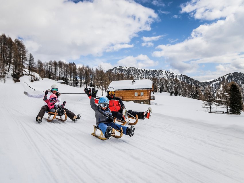 Geheimtipp Hotels in Südtirol 😎☀️ Familie rodelnd im Schnee vor einer Berghütte und bewaldeten Bergen