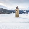 Die Entstehung eines altbekannten Brauches Kirchturm ragt aus zugefrorenem See mit schneebedeckten Bergen im Hintergrund